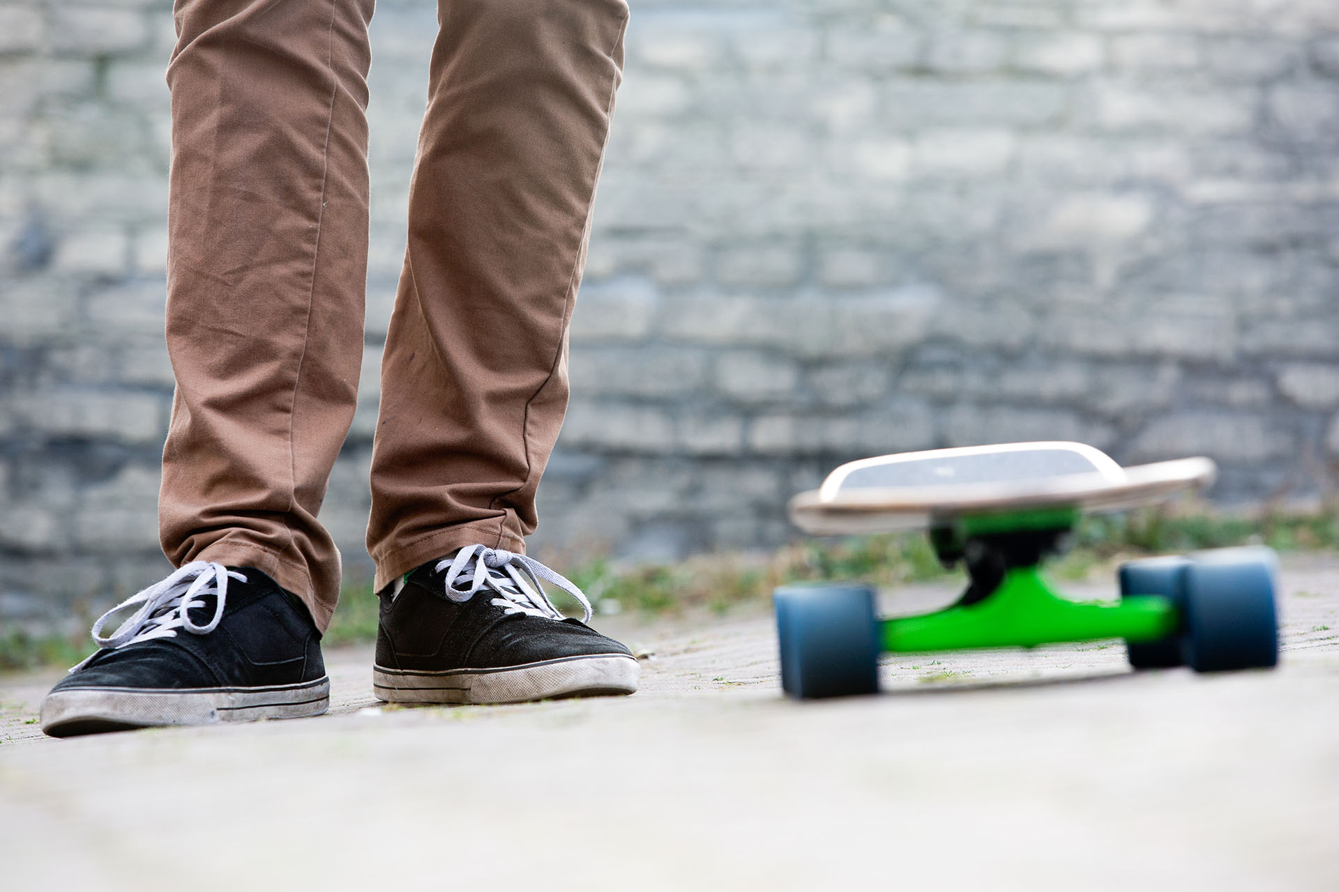 The two feet of a casually dressed man standing next to a skateboard in an urban setting, with a brick wall in the background. All earthy tones